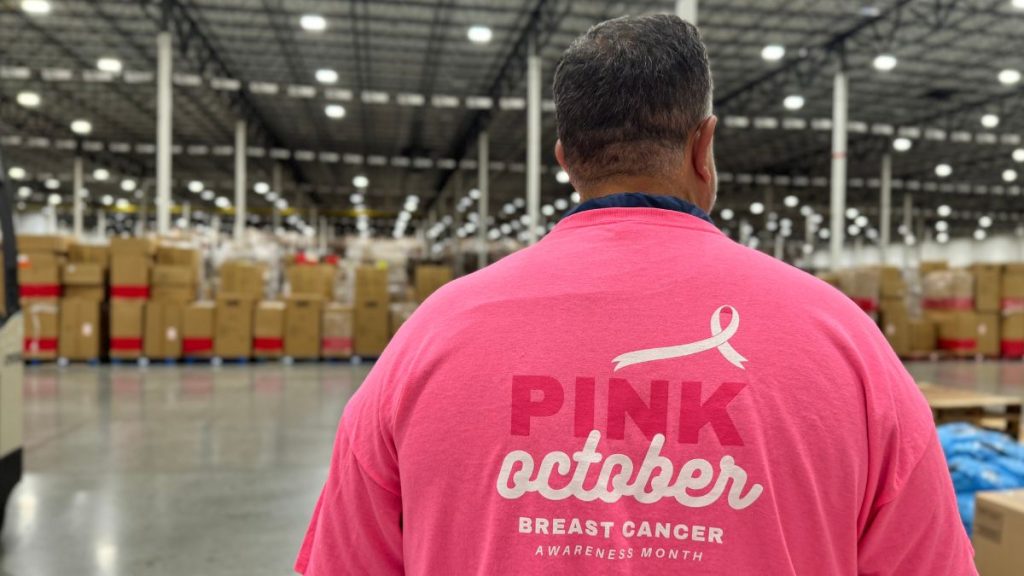 Person wearing a pink 'Pink October' Breast Cancer Awareness shirt, seen from behind in a large warehouse with boxes in the background.