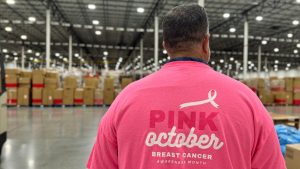 Taylor Logistics is turning pink for Breast Cancer Awareness Month - Taylor Logistics Inc. Person wearing a pink 'Pink October' Breast Cancer Awareness shirt, seen from behind in a large warehouse with boxes in the background.